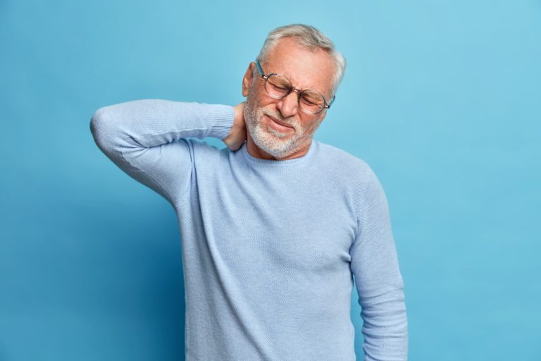 Homme âgé en douleur, main sur la nuque, exprimant une gêne cervicale sur fond bleu uni